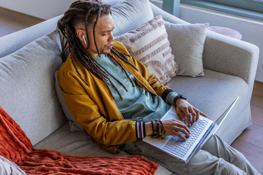 African American man typing on laptop on sofa at home wearing mustard jacket, white earbuds