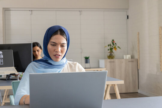 Female coworkers working in shared office with woman in blue headscarf using silver laptop