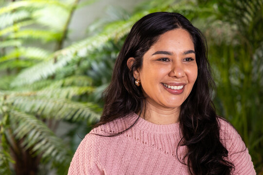 Indian woman late twenties smiling, looking off-camera in garden with pink knit sweater, gold hoops