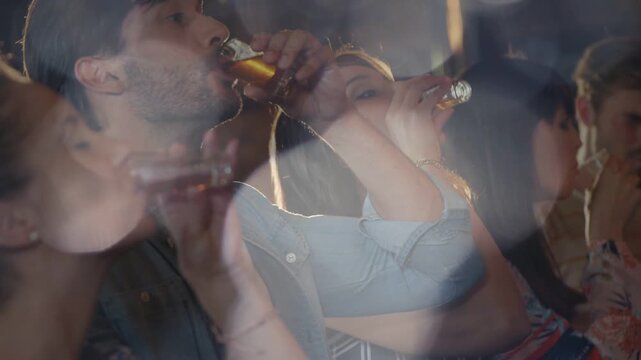 Man in denim initiating toast raising shot glass, group drinking amber shots amid ghosted bar bokeh