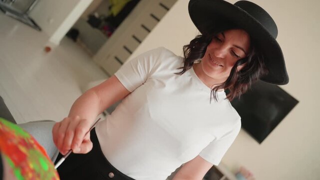 Pregnant woman smiling in wide brim hat, gently touching colorful wrapping paper on table in bright apartment, candid lifestyle shot showing warm domestic preparation for baby, soft lighting
