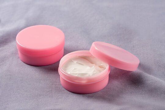 Open pink cream jar with smooth white cream , one opened the smooth texture on soft purple fabric, for beauty, skincare, and facial treatment product.