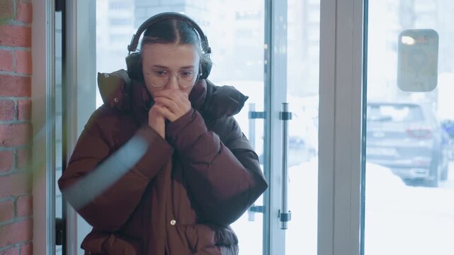 White commuter steps into cafe, exhaling cold breath and settling hands, headphones and heavy coat against brick wall, plant foreground and frosted glass, calm content expression in cozy interior