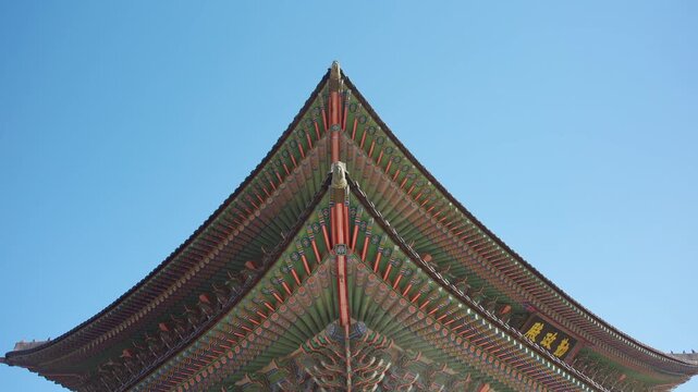 Ornate tiered roof of Gyeongbokgung Palace rises against a clear blue sky, showcasing vibrant dancheong patterns in green, red, and gold with traditional Korean wooden eaves and carved details.