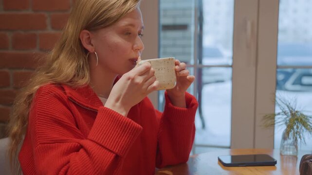 Barista serving coffee at cafe, carrying ceramic mugs to table, customer in red sweater receives cup, window seating with urban street view, apron and tray visible, brief conversation and warm morning