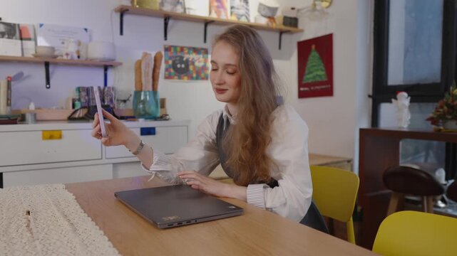 Charming young caucasian woman sitting at table in a modern cafe, smiling while having a video call or taking a selfie with her phone before starting to work on her laptop