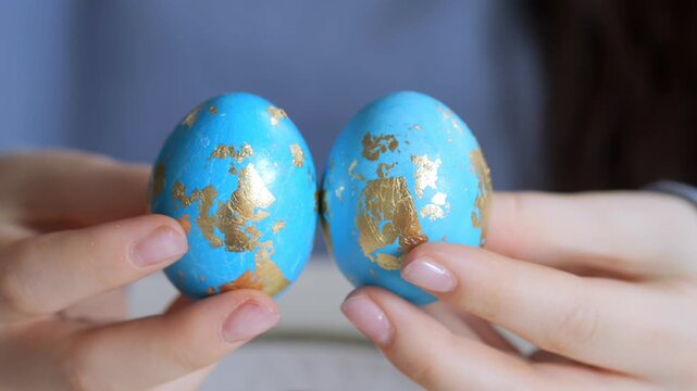 Hands hold two blue eggs with gold leaf patterns. Eggs appear handcrafted, festive, and decorative. Focused close-up highlights texture and color