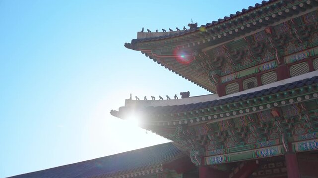 Golden sunlight peeks through ornate traditional Korean palace rooftops featuring intricate painted wooden eaves and decorative roof guardians against a clear blue sky at Gyeongbokgung Palace in.