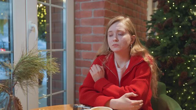 White woman alone at cafe with arms crossed in red sweater by brick wall and Christmas tree, empty cup on table, winter light through window, resigned expression and quiet holiday isolation
