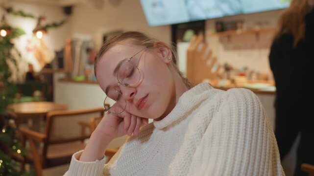 White student napping in busy cafe, festive evening scene with staff moving behind counter and twinkling tree lights white student in chunky sweater and glasses leans head on hand during study break,