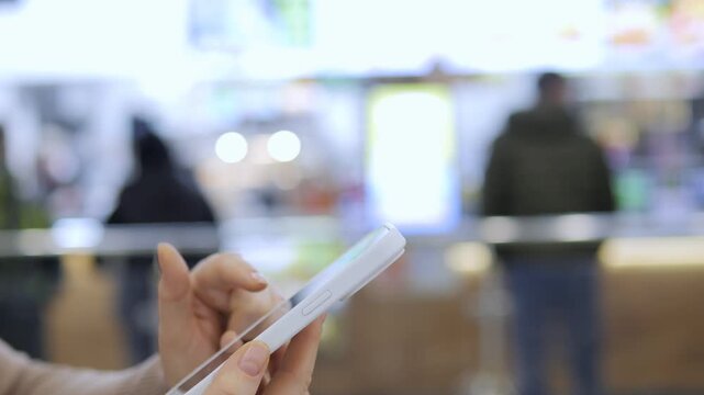 Hand holds white smartphone in public space. Background shows blurred people and bright lights. Focus remains on device and hand only. No animals or plants visible