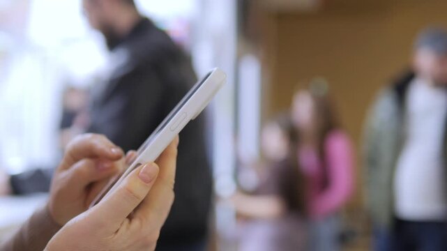 Close-up of hands using a smartphone. Background shows blurred people in indoor space. Focus remains on device and fingers interacting. Lighting is soft, natural, and evenly distributed