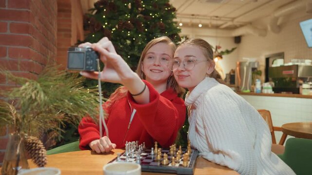 Two white friends in cafe selfie wearing festive sweaters, leaning close over chessboard and coffee, pointing camera for group portrait, warm Christmas tree lights and brick interior, candid laughter