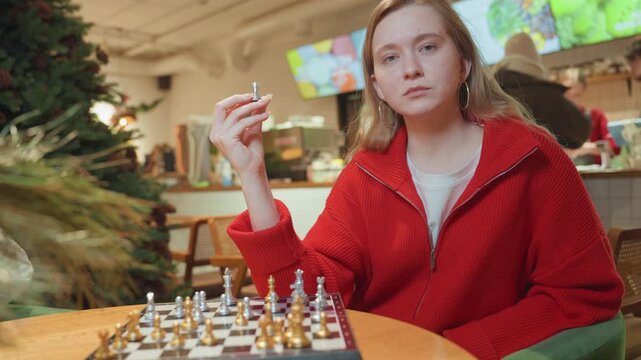 White woman in cafe making decisive move on chessboard, red sweater, intense expression, ambient holiday lights, menu screens and patrons in background, focused hand motion, modern coffeehouse vibe