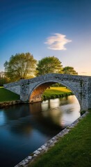 Obraz premium An elegant stone bridge arches over a serene flowing river surrounded by lush green trees and blue skies in a quiet and natural environment, arch, majestic, blue