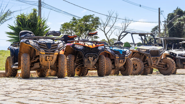 A row of muddy ATVs and UTVs are parked on a cobblestone road with trees and power lines in the background.