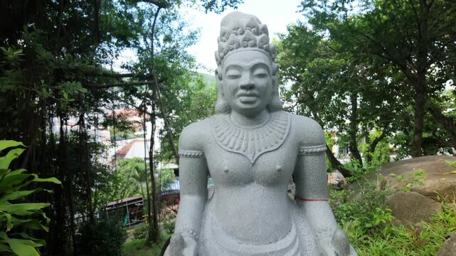 A stone statue of a seated Buddha is displayed outdoors in a lush green garden setting in Po Nagar Temple, Nha Trang, Vietnam.