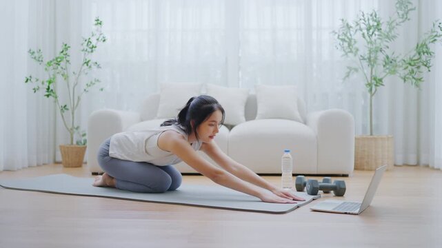 Young Asian woman stretching yoga workout on exercise mat while online training class with computer laptop in living room