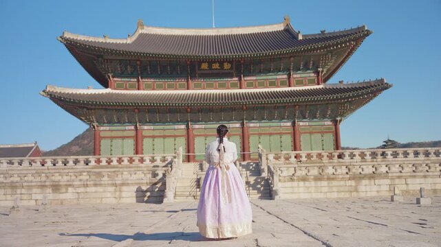 Young woman in elegant pink and white traditional Korean hanbok dress admires the ornate Gyeongbokgung Palace throne hall under clear blue skies in Seoul, with mountains visible in the distance.