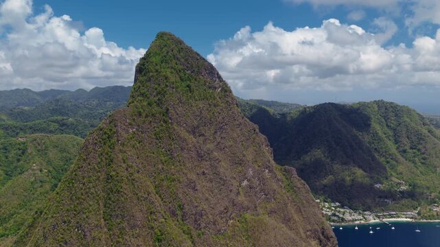 Aerial drone footage captures majestic Piton volcanic peak towering over lush green rainforest mountains in Saint Lucia. Turquoise Caribbean bay with boats and coastal village below cloudy blue sky.
