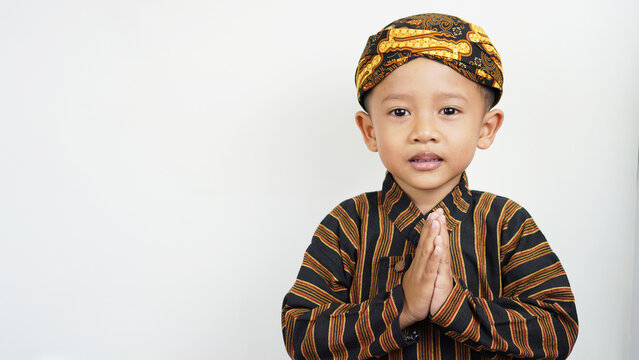 Portrait of a cute and handsome Asian boy of preschool age, wearing traditional East Javanese clothing, namely Baju Lurik complete with his blangkon while greeting and smiling sweetly,