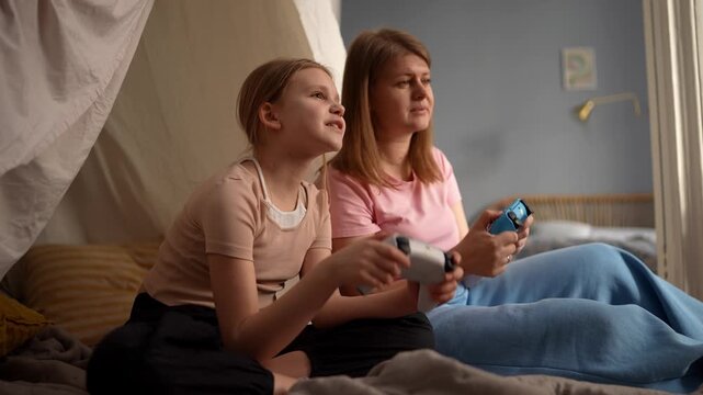 Two girls playing game controllers, soft daylight through window, bed and pillows as backdrop, relaxed smiles and gentle banter, cooperative play with shared .