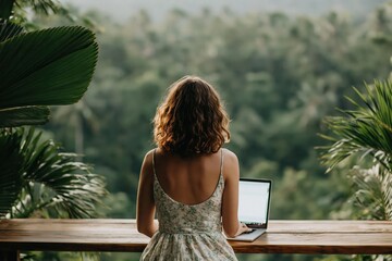 Obraz premium Woman in dress works on her laptop on wooden balcony surrounded by lush green plants with misty jungle landscape in background. High quality photo