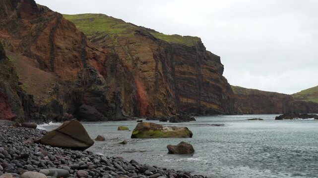 Wide shot of a dramatic, rugged coastal landscape featuring a pebble beach, calm ocean waves, and impressive stratified cliffs under a cloudy sky. Sao Lorenco, Madeira, Portugal