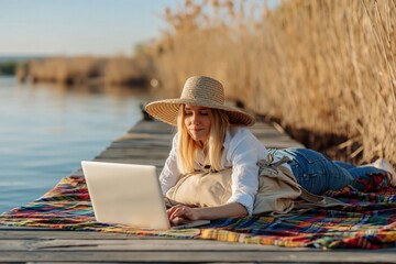 Naklejka premium Woman in wide-brimmed hat lies on wooden dock, engrossed in her laptop. Scene is bathed in warm light of sunset, with calm water and dry reeds in background. High quality photo