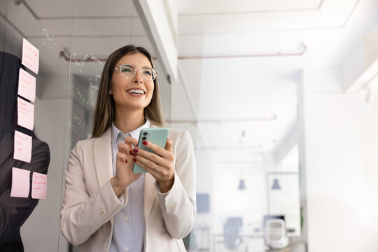 Laughing businesswoman using smartphone in coworking, stands next to wall with attached sticky notes engaged in planning, management, exchanging messages at workplace. Innovative business tech suage