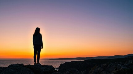 Obraz premium A person is standing on rocks by the sea during sunset. The sky shows bright colors of orange and blue. The scene captures the beauty of nature.
