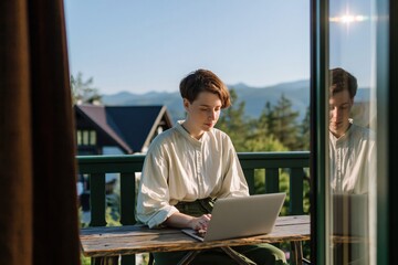 Obraz premium Woman is focused on her laptop while sitting on wooden balcony, surrounded by beautiful mountain landscape and clear blue sky. High quality photo
