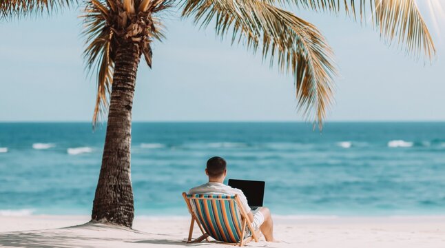 Man sits in beach chair on white sand beach, working on his laptop. Palm tree provides shade as he enjoys ocean view. High quality photo