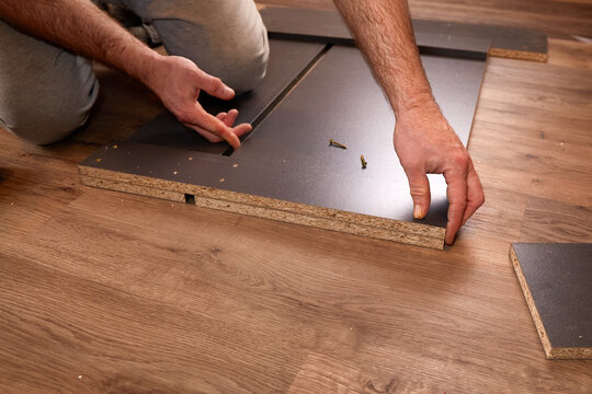 Man assembling furniture on wooden floor, connecting panels with screws.