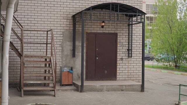 Urban apartment back entrance metal canopy, rusty brown door under black metal awning, exterior metal staircase, wooden trash container with blue liner, gravel pavement, pale brick facade, single