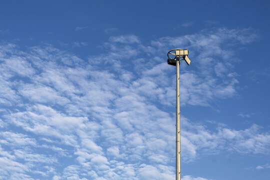 ​A tall metal high mast lighting tower equipped with LED floodlight fixtures, set against a beautiful blue sky filled with white altocumulus clouds.