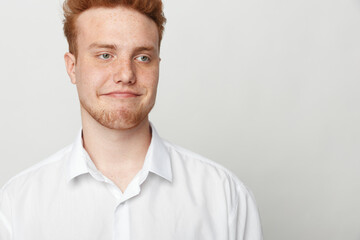 Obraz premium Young redhead man in white shirt looks sideways with skeptical expression on light background. Concept of doubt, uncertainty, and casual portrait of male model with freckles.