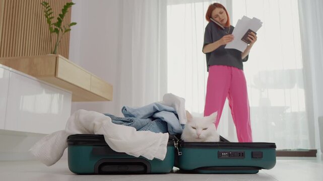 A cat sits inside a suitcase filled with clothes. A woman stands nearby with her passport and papers, talking on the phone about travel plans and issues.