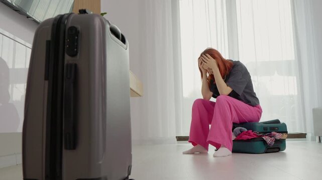 A woman sits on a suitcase with clothes inside while holding her head in her hands. She looks stressed as she thinks about packing for her trip.