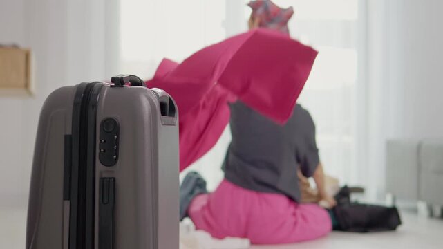 A woman is unpacking clothes from a suitcase. She throws clothes behind her while sitting on the floor in a bright room filled with natural light.