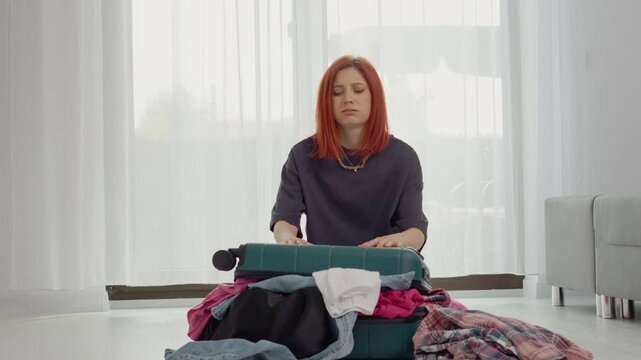 A woman sits on the floor and leans on her suitcase. She looks worried as she tries to close her suitcase full of clothes for her trip.