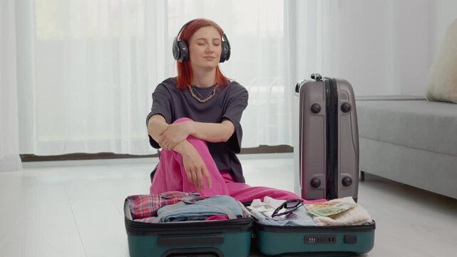 A woman sits in a room with her suitcase open. She listens to music through headphones. Clothes and money in euros are nearby. She looks relaxed and lost in thought.