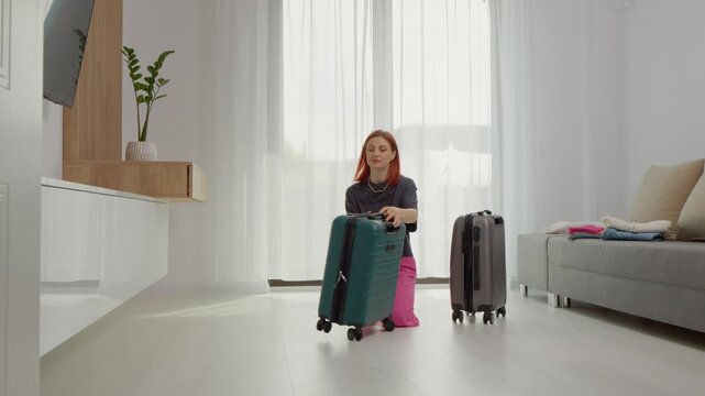 Woman stands in living room and opens two empty suitcases as she prepares for a trip. She checks the bags while arranging items around her in the space.