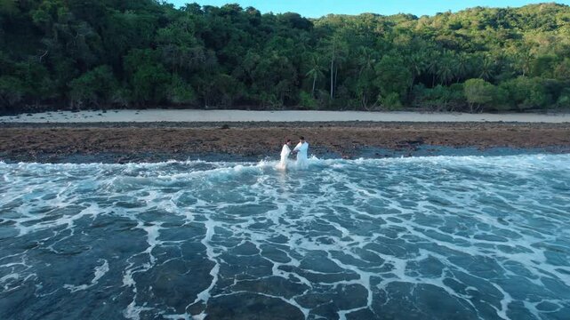 Aerial view moving backwards from an Indonesian couple in white to reveal the rocky shore, crashing ocean waves, and lush tropical forest background.
