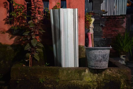 A black plastic bucket sits on the edge of a concrete decomposition pit containing soil and plants, located next to a red wall in a Balinese shrine area under evening artificial light.