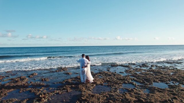 Drone shot flying backward from an Indonesian couple standing on a rocky beach, looking at the blue sea during golden hour sunset.