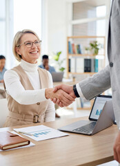 Fototapeta premium Smiling mature businesswoman with glasses shaking hands with a male colleague over a desk with a laptop and financial charts in a bright modern office.