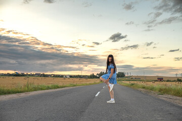 Fototapeta premium Young woman in blue floral dress stands on dirt road beside golden wheat field at sunset, with distant houses and cloudy sky in the background