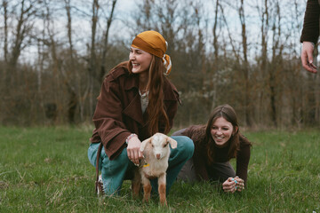 Fototapeta premium Two cheerful girls pet a curious goat kid in a grassy field, enjoying a carefree outdoor moment, natural smiles, warm clothing, gentle breeze, countryside vibes, and a playful, friendly mood.