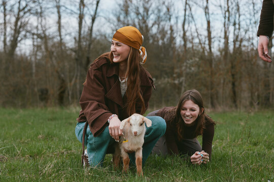 Two cheerful girls pet a curious goat kid in a grassy field, enjoying a carefree outdoor moment, natural smiles, warm clothing, gentle breeze, countryside vibes, and a playful, friendly mood.